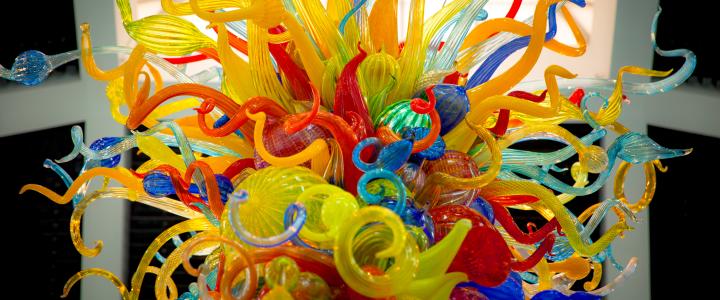 Vibrant glass sculpture by artist Dale Chihuly, with twisting, multicolored tendrils and abstract shapes, featuring red, yellow, blue, and green hues against a neutral background.