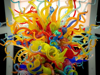 A vibrant, intricate glass sculpture with swirling shapes in yellow, orange, red, blue, and green hues set against a lit background.