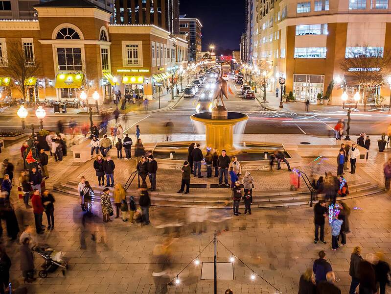 A busy Town Center at night with a central fountain, surrounded by people walking and socializing. Buildings and streetlights illuminate the scene.