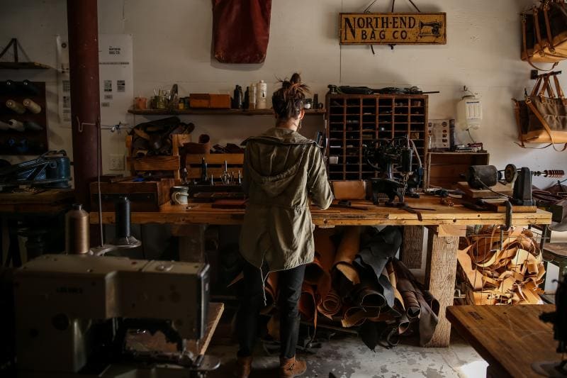 Person stands at a cluttered workbench in a leather workshop, surrounded by tools and materials, with a "North End Bag Co" sign above.