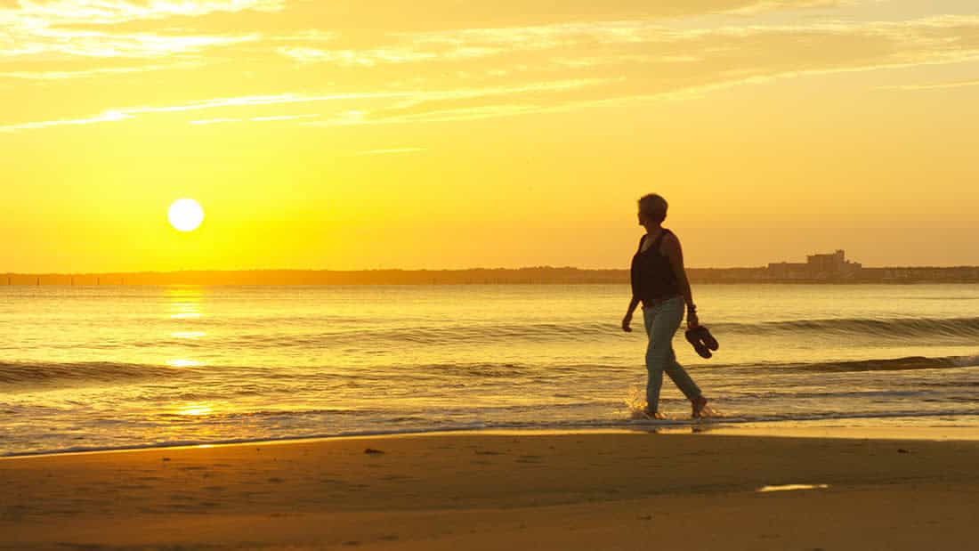 Person walking along Virginia Beach at sunset, holding sandals, with calm ocean waves and an orange sky.