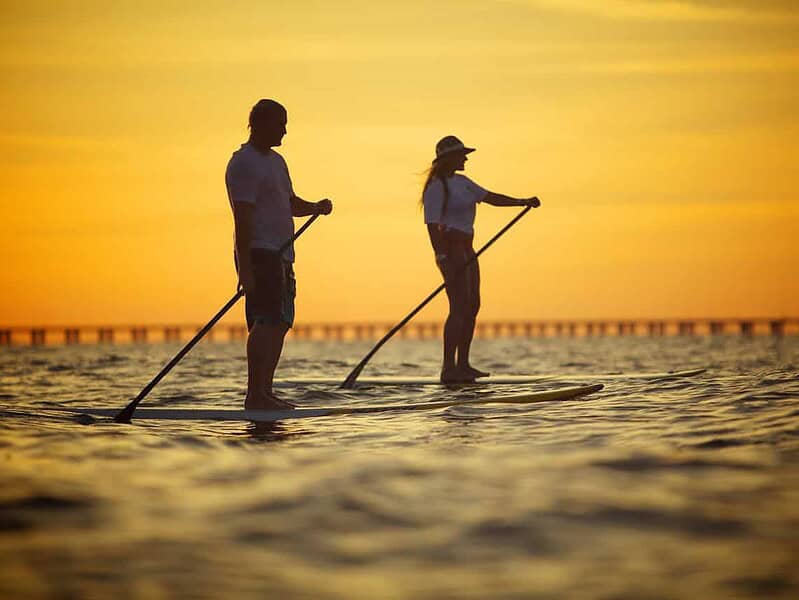 Two people are stand-up paddleboarding on calm water at sunset, with a bridge visible in the background.