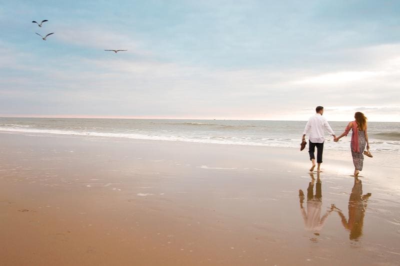 A couple walks barefoot along a beach holding hands, with gentle waves and birds in the sky on a cloudy day.