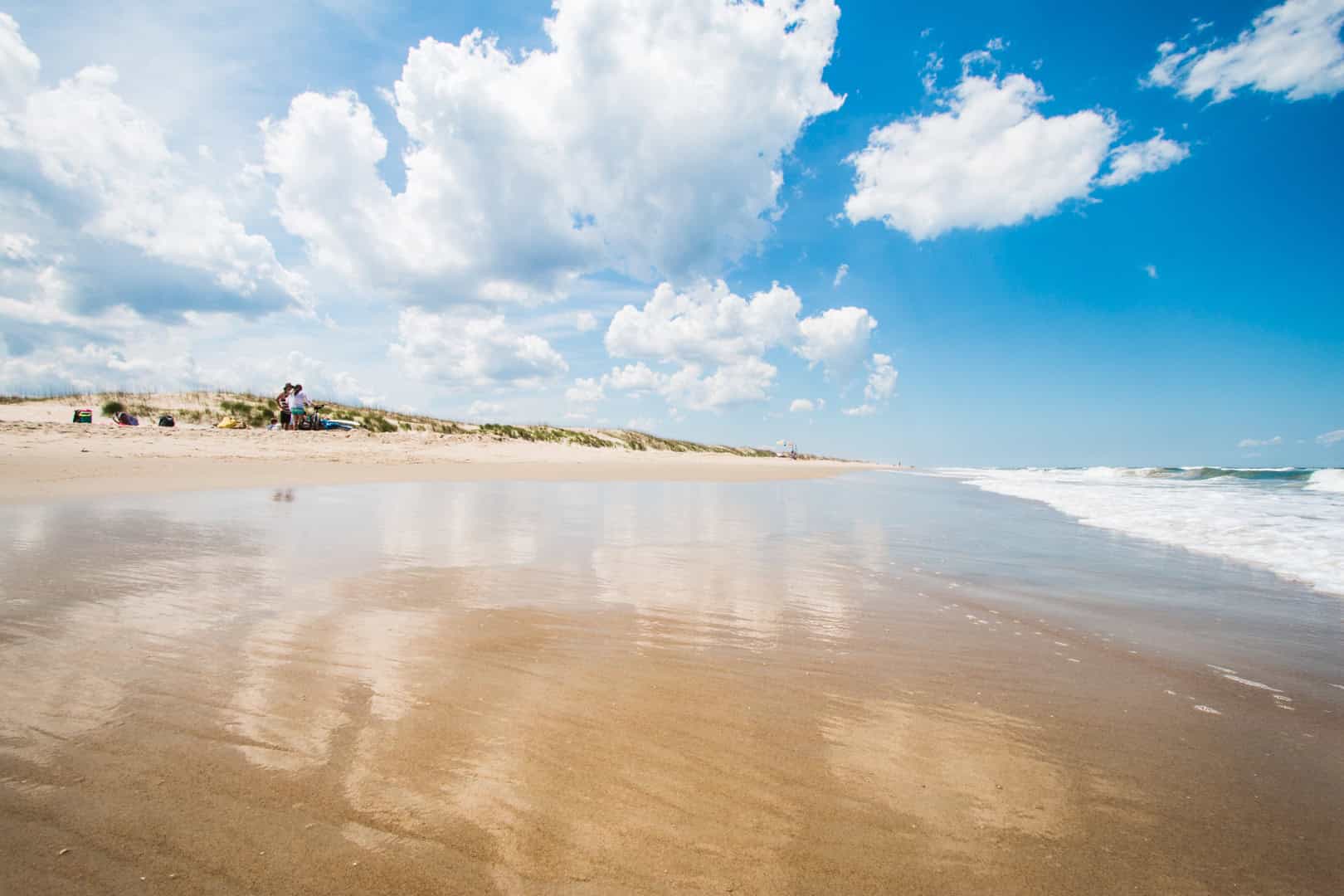 Sunny beach scene with wet sand reflecting the sky, scattered clouds, and rolling waves. Sparse vegetation on a dune in the background.