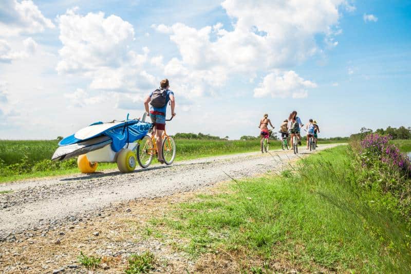 Four people ride bicycles down a gravel path. One pulls a trailer with surfboards. The path is bordered by grass, and the sky is partly cloudy.