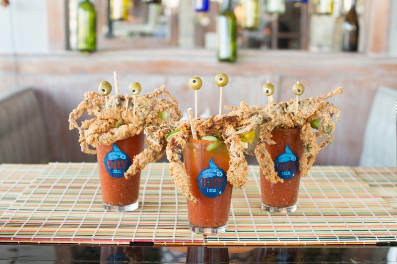 Three Bloody Mary cocktails garnished with fried soft-shell crabs, lime slices, and olive-topped skewers, set on a colorful table mat.