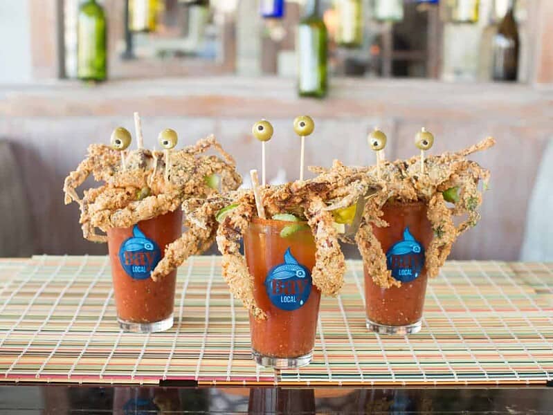 Three Bloody Mary cocktails at Bay Local with fried soft shell crab garnishes, lime wedges, and green olive skewers on a bamboo mat.