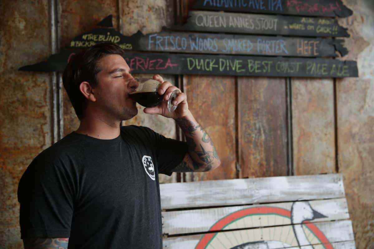 A man with tattoos drinks a dark beer from a glass, standing near a rustic wall with wooden signs listing various beer names.