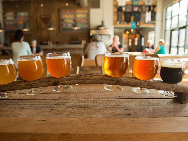 A row of six beer glasses filled with different types of beer on a wooden tray in a brewery setting.