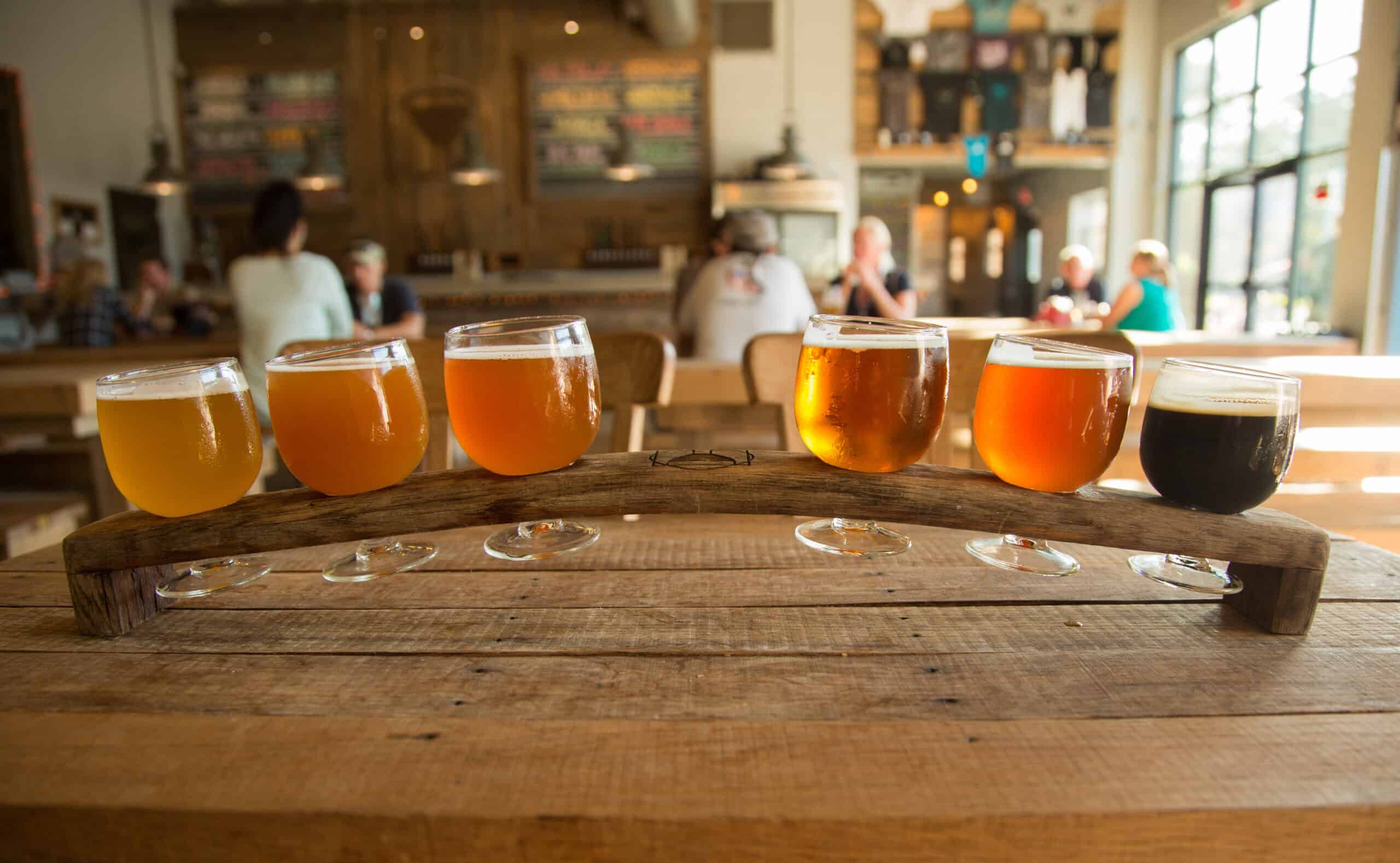 A row of six beer glasses filled with different types of beer on a wooden tray in a brewery setting.