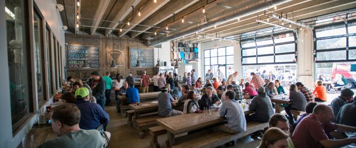 People sitting at long wooden tables in a bustling brewery taproom with high ceilings, chalkboard menus, and large windows.