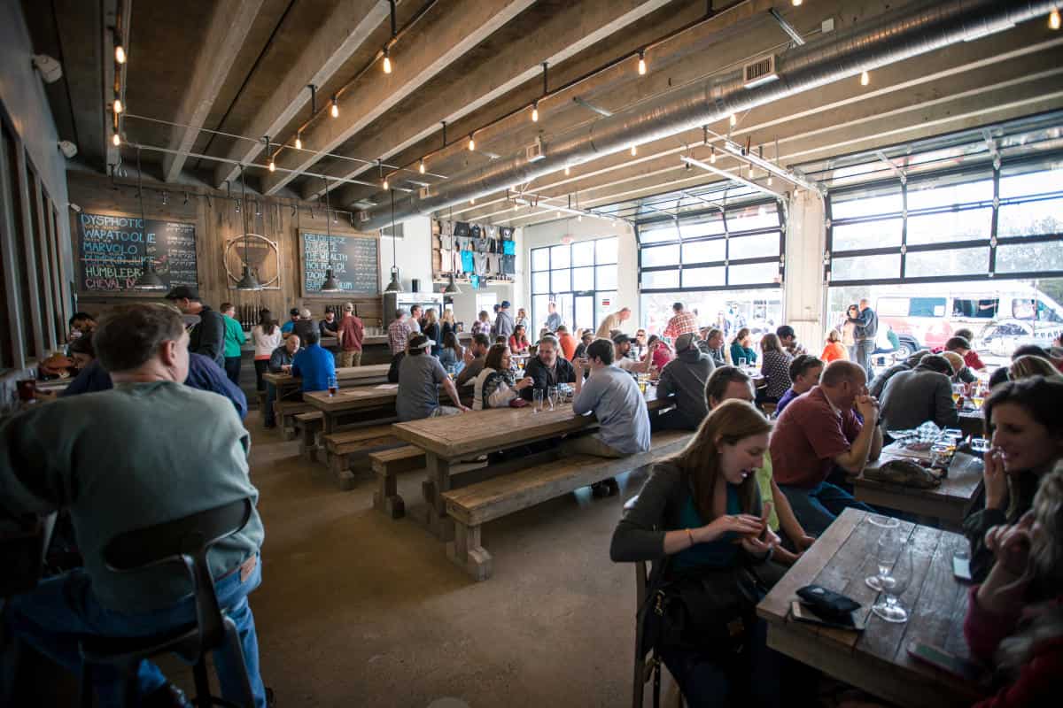 People are sitting at communal wooden tables in a bustling industrial-style restaurant with large windows and exposed beams.