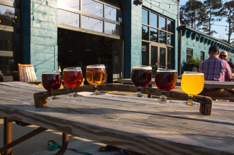 A wooden paddle holds six glasses of assorted craft beer on an outdoor wooden table. Blue building and people in the background.