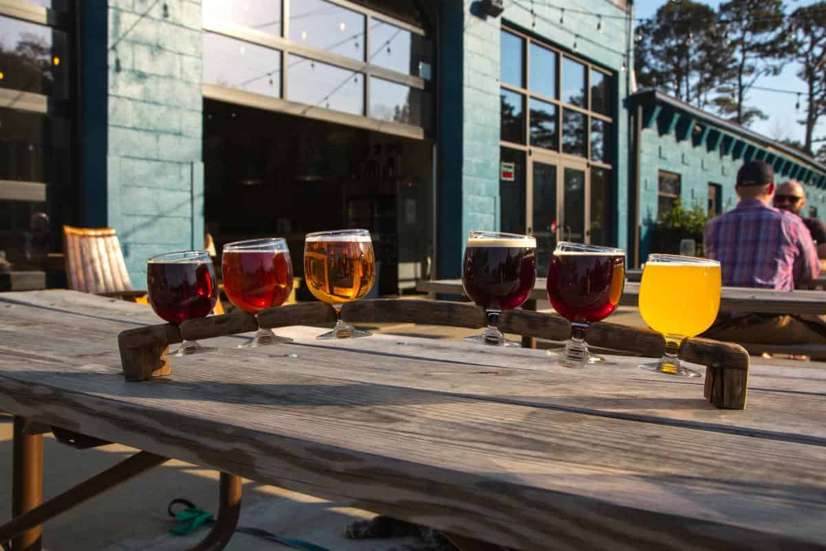 A flight of six beers with varying colors sits on a wooden table outdoors, near a teal-blue building under a sunny sky.