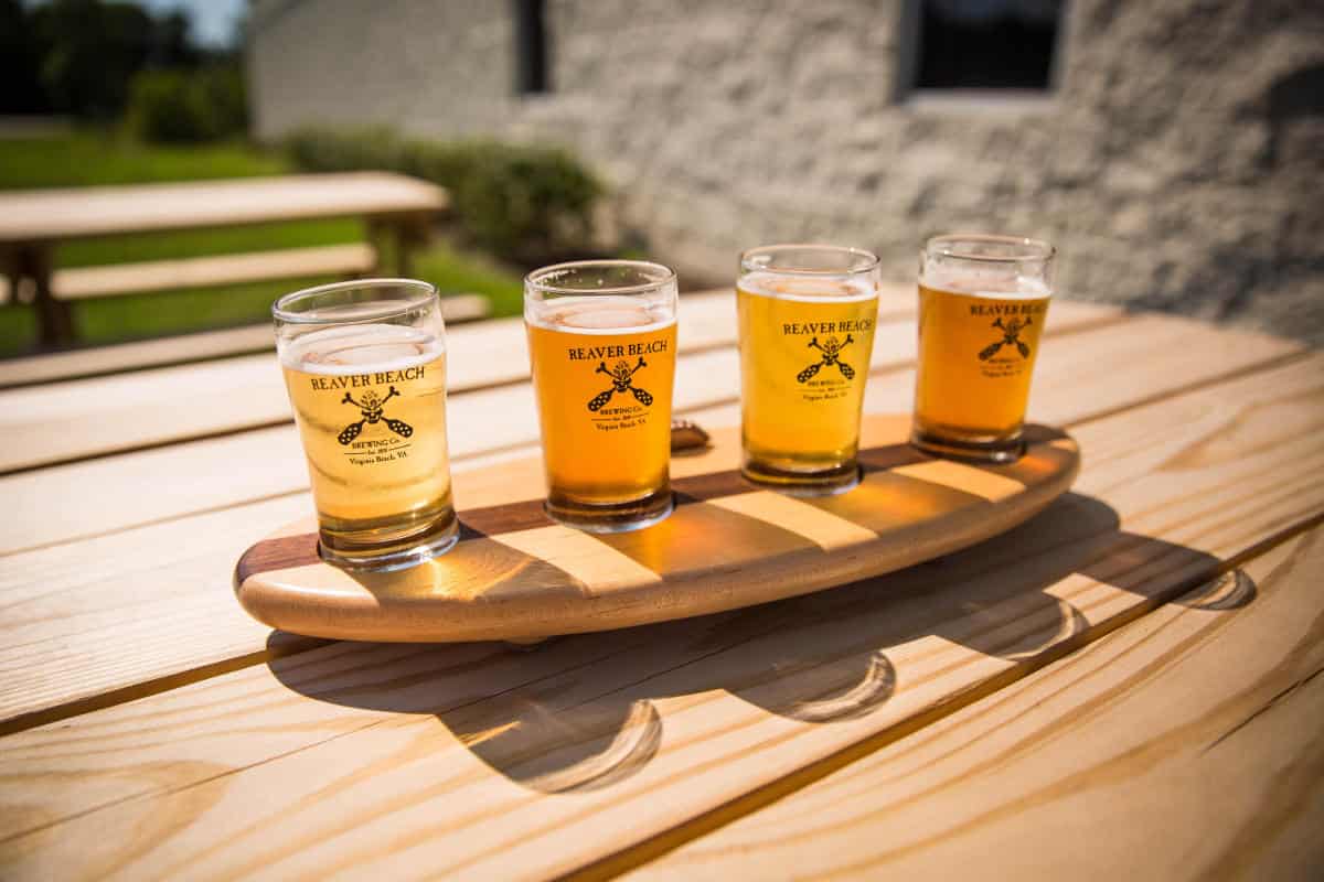 Four beer samples in branded glasses on a wooden paddle, placed on an outdoor wooden table with a building in the background.