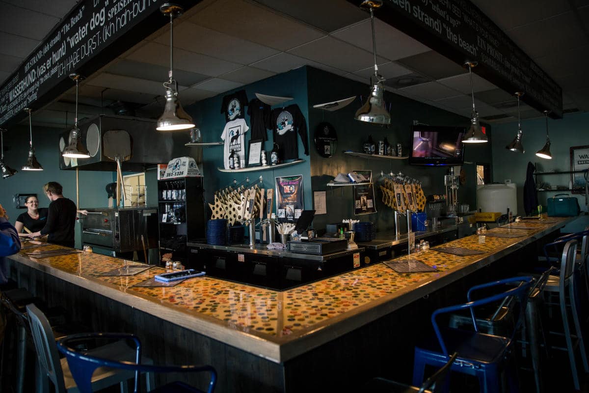Interior of a bar with a U-shaped counter, stools, hanging lights, and various decorations on the walls. Two people stand near the bar area.