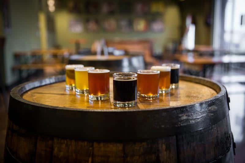A variety of seven beer samples arranged on a wooden barrel in a dimly lit bar with wooden tables and chairs in the background.