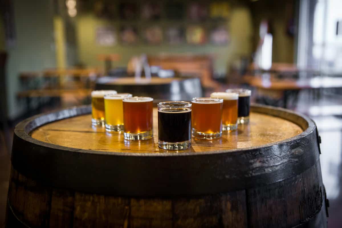 A variety of beer samples in small glasses are arranged on a wooden barrel in a rustic setting.