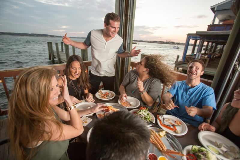 Group of people laughing and enjoying a meal at a waterfront restaurant. Plates of seafood and salads are on the table.