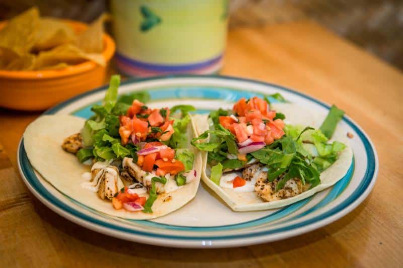 Two tacos with grilled chicken, lettuce, tomatoes, and onions on a plate. A bowl of tortilla chips and a colorful mug are in the background.