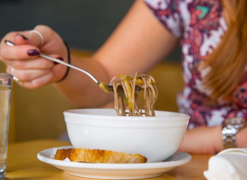 Person holding a fork with noodles above a white bowl. A slice of bread is on the side, and there's a glass of water nearby.