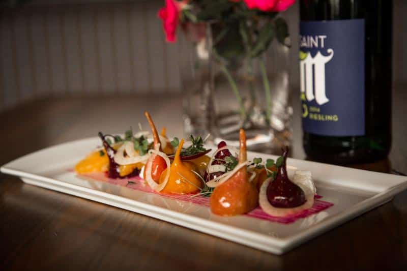 A gourmet dish of colorful beetroot portions arranged on a rectangular plate, garnished with greens and onions, next to a wine bottle and a vase with pink flowers.