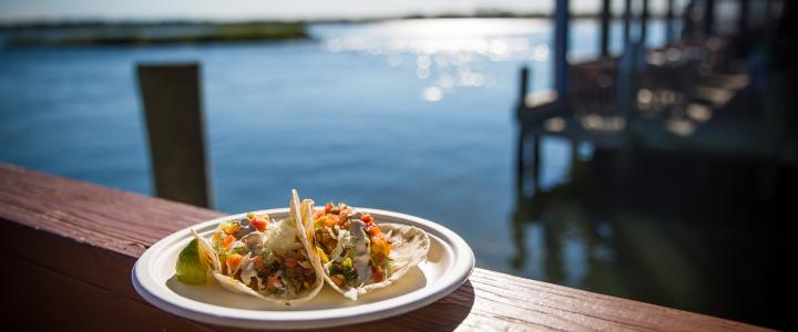 Two tacos filled with vegetables and salsa on a plate, placed on a wooden railing by the water with a dock in the background.