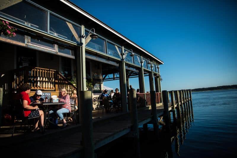 People seated at a wooden deck restaurant by the water under a clear blue sky.