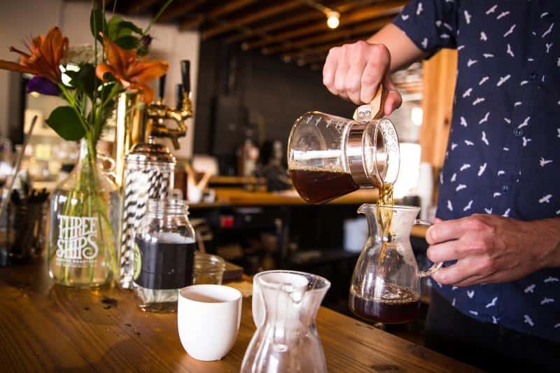 Person pouring coffee from a glass pot into a small carafe on a wooden counter, with a vase of flowers and a white mug nearby.