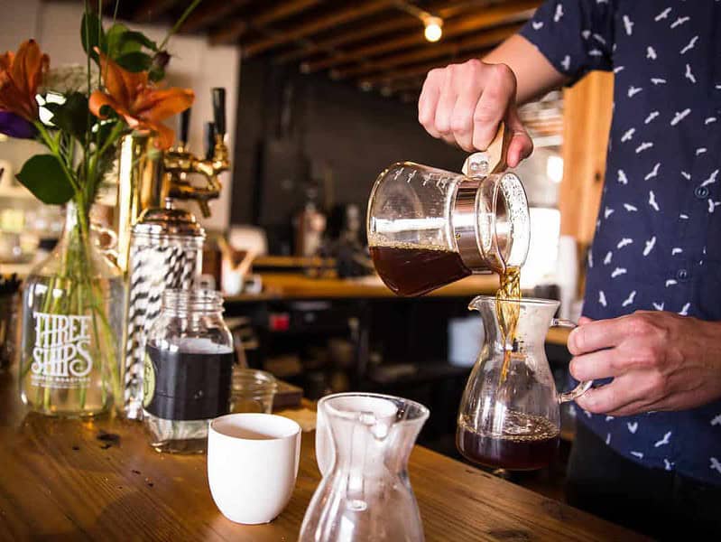 A person pours coffee from a glass pitcher into another on a wooden counter. Fresh flowers and coffee equipment are nearby.