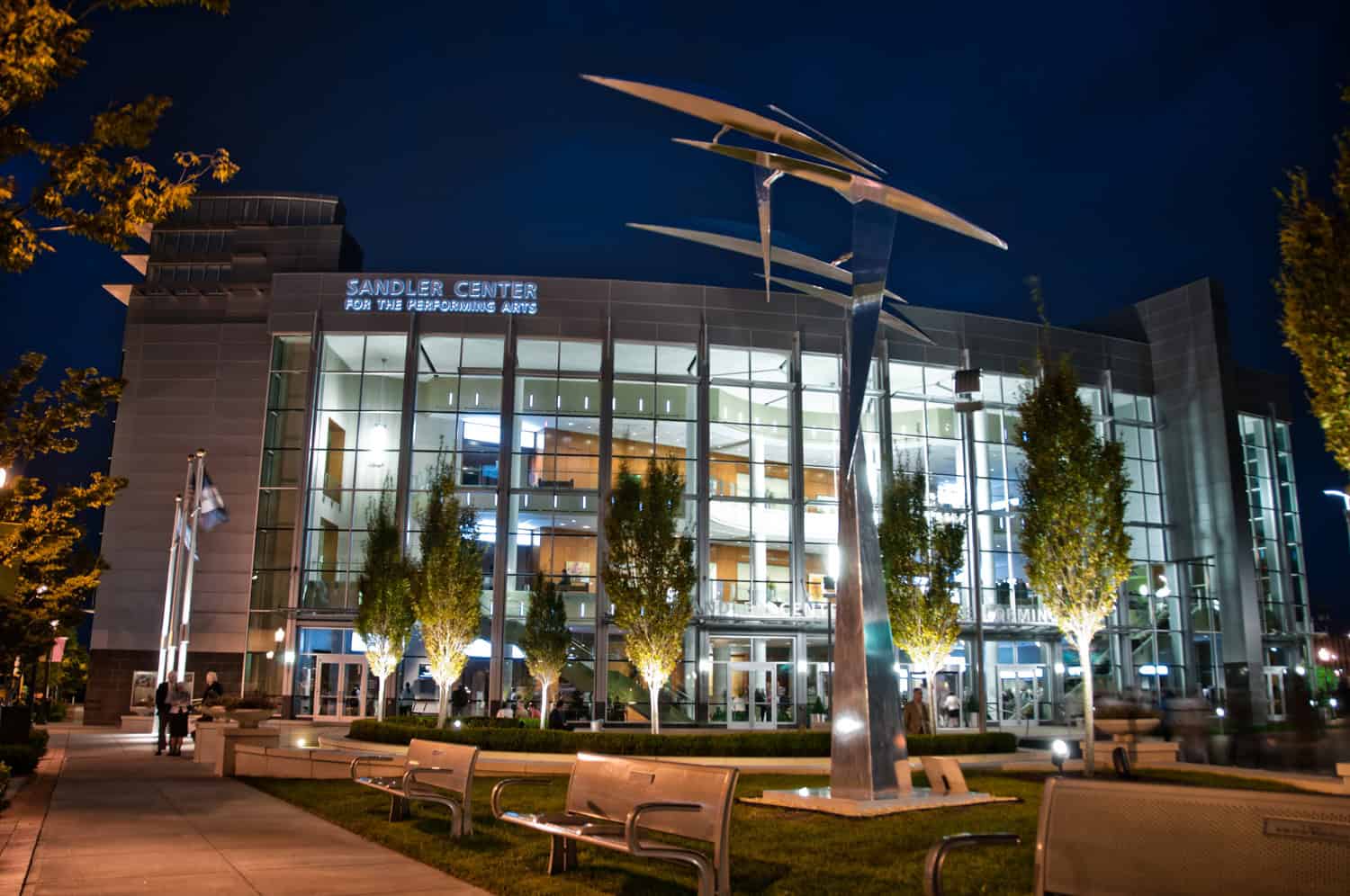 Exterior of Sandler Center for the Performing Arts at night, with illuminated glass facade and a large metal sculpture in front.
