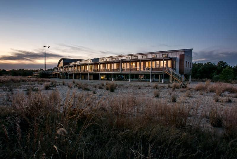 A modern, rectangular building with a raised platform stands in a grassy field at dusk. The lights inside are on, and the sky is clear with a few clouds.