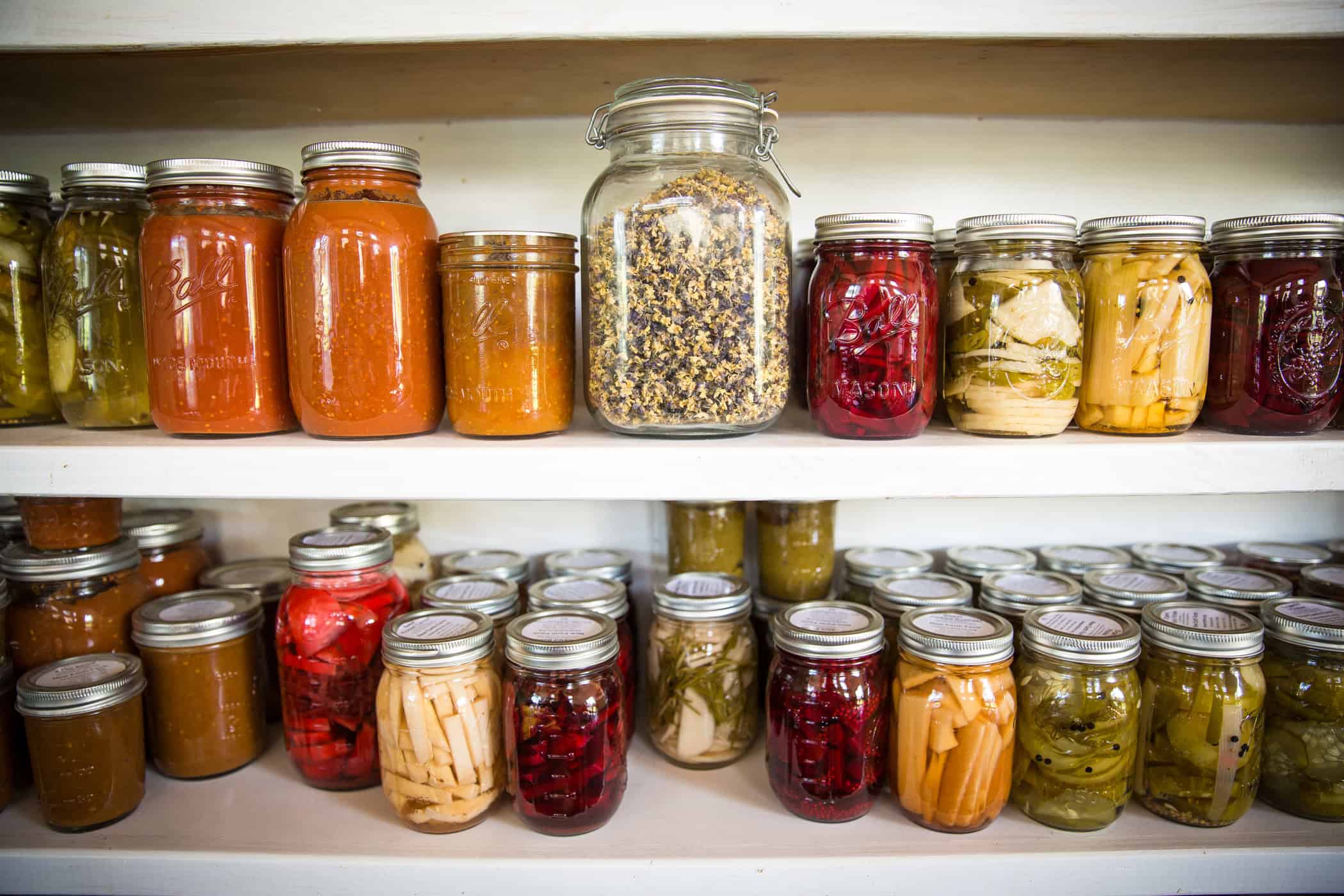 Jars of various preserved foods, including sauces, pickles, and dried herbs, arranged on white shelves.