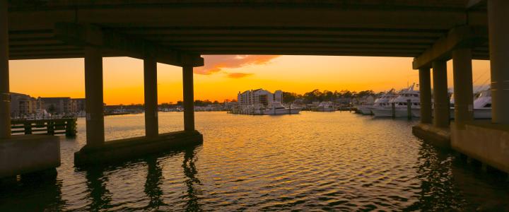 Under a bridge, a marina with boats is visible at sunset. The water reflects the orange and pink hues of the sky.