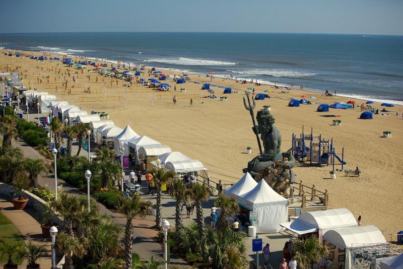 Beach scene with tents and people along the shoreline. A large statue of Neptune holding a trident is visible. The ocean is calm under a clear blue sky.