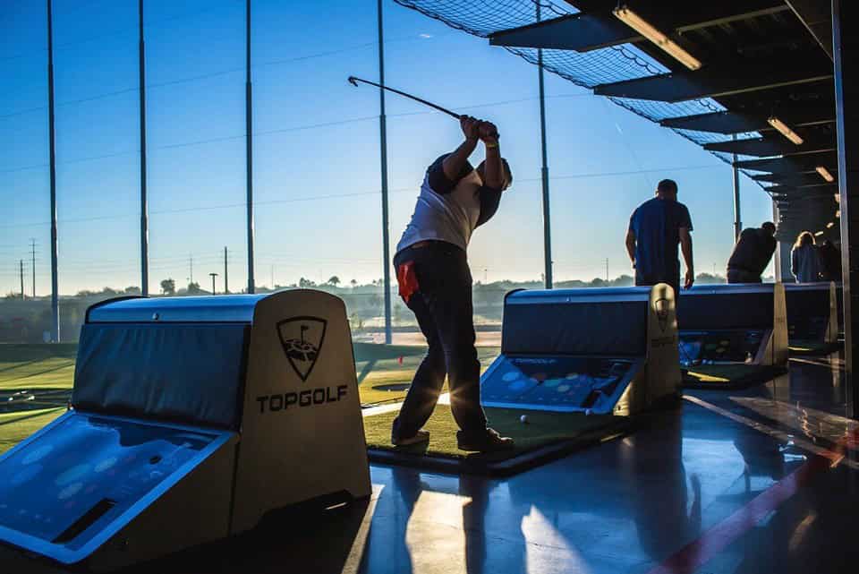 A person swings a golf club at an indoor driving range, silhouetted against a large window with several golfing bays visible.