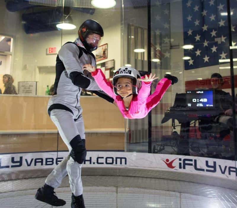 An instructor assists a child wearing pink in an indoor skydiving wind tunnel.