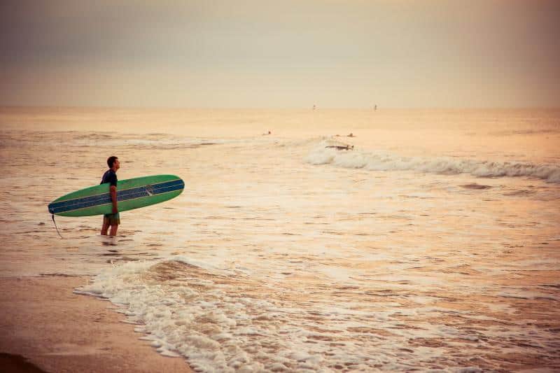Person holding a surfboard stands in shallow water at the beach during sunset; small waves roll in the background.