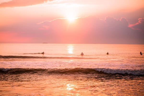 Surfers in the ocean under a pink and orange sky, with the sun setting behind clouds, reflecting on the water.