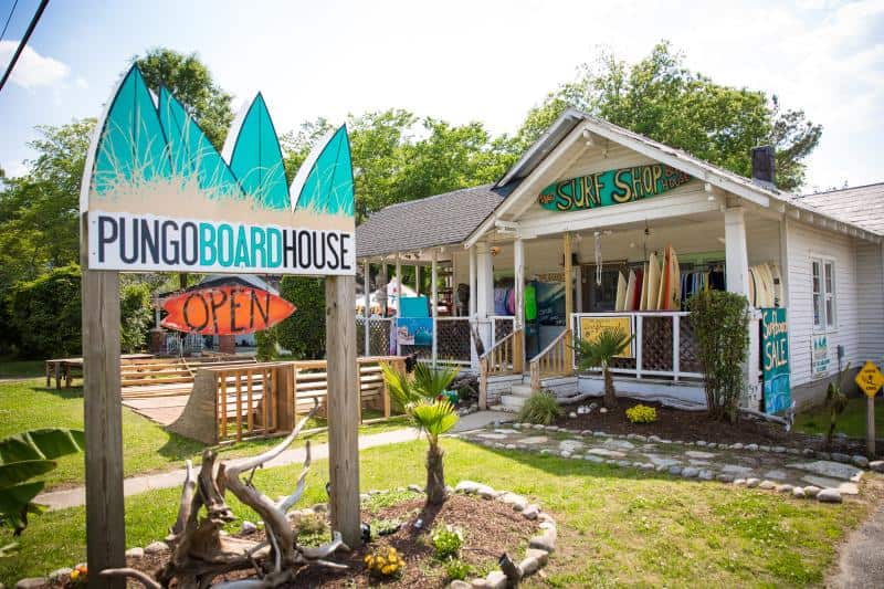 Surf shop with surfboards and tropical decor outside, located in a small building with a sign reading "Pungo Boardhouse." The area is surrounded by grass and trees.