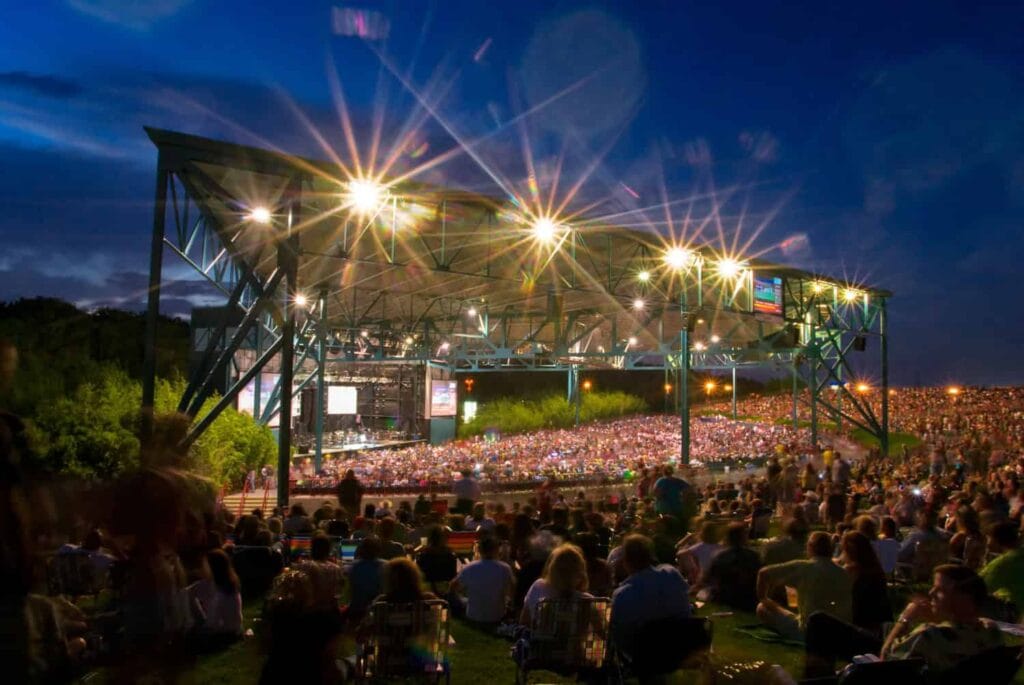 A large crowd gathers on a grassy hill under a starry sky, facing an illuminated outdoor concert stage with bright lights.