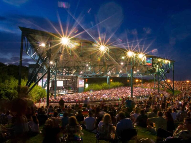 The Veteran's United Home Loans Amphitheater is lit up during a nighttime concert, with a large audience seated on the lawn and under the pavilion.