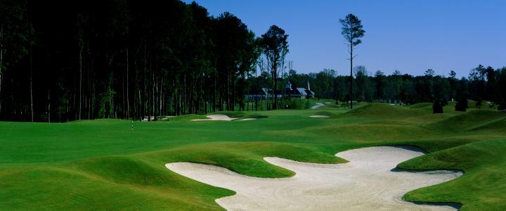 Golf course landscape with green fairways, sand bunkers, and surrounding trees under a clear blue sky.