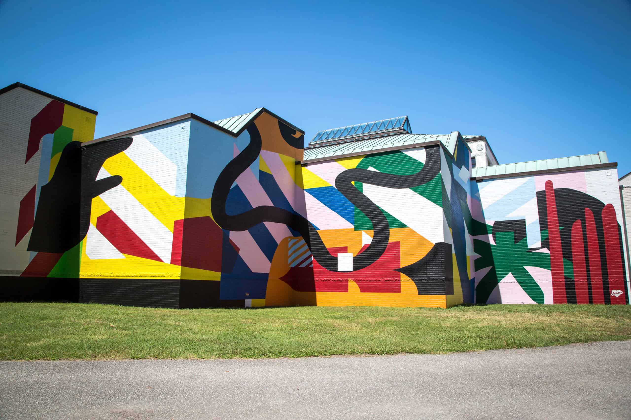 Colorful abstract mural covers building exteriors, featuring geometric shapes, wavy lines, and bold colors like red, yellow, black, and green under a clear blue sky.