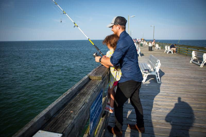 A man helps a young girl fish off a wooden pier on a sunny day. Several people and benches are visible in the background.
