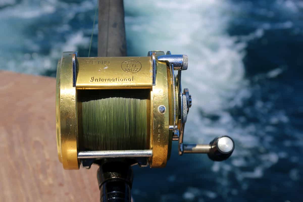 Close-up of a gold fishing reel attached to a rod, with green line spooled. The background shows blurred water with white foam.