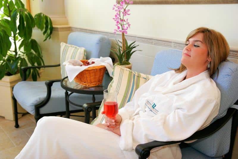 A person in a white robe relaxes on a cushioned chair, holding a glass of red beverage. A basket of towels and a potted plant are in the background.