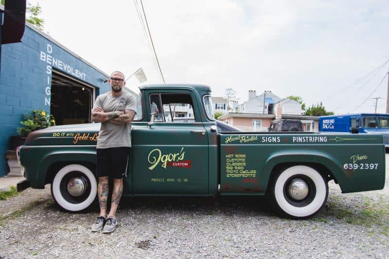 Man with tattoos stands next to a vintage green truck with custom signage outside a shop.