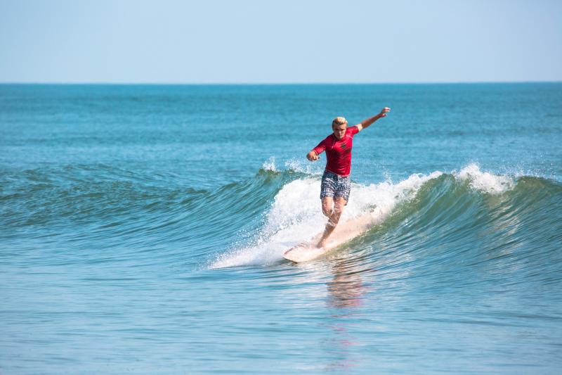 A person in a red shirt and shorts surfs on a small wave in the ocean under a clear blue sky.
