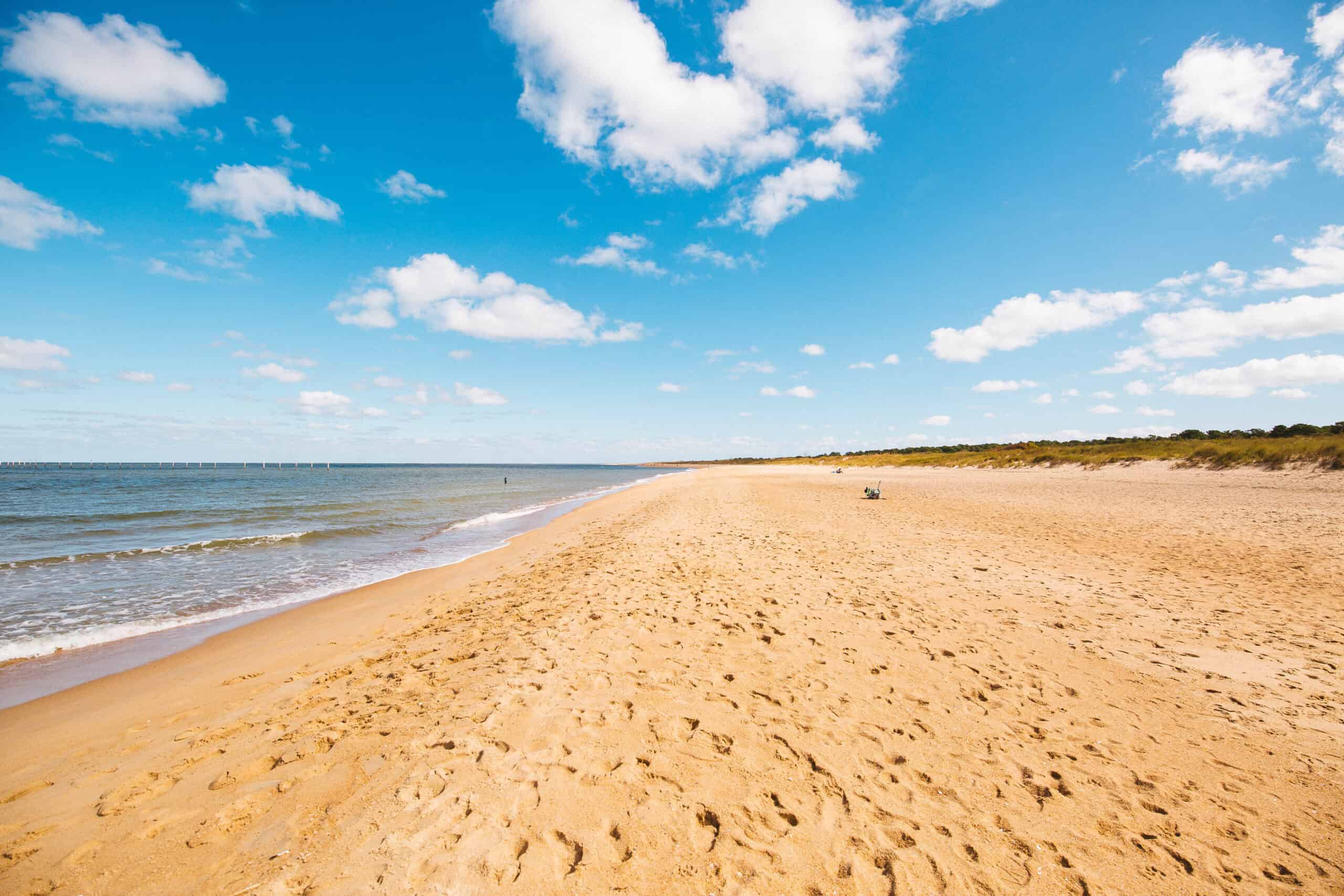 Sandy beach with gentle waves under a blue sky scattered with clouds. A single person sits on the sand, and greenery lines the far right edge.